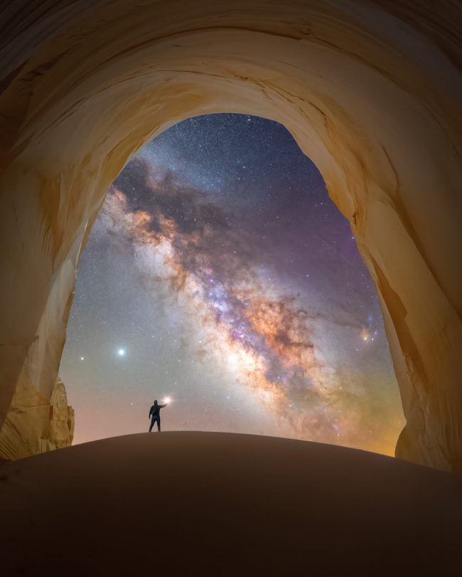 Man standing under rocky arch, pointing at a star or planet, with Milky Way cutting through sky behind.