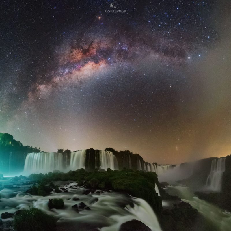 Wide waterfalls in foreground with arching Milky Way behind and other light effects in the sky.