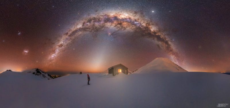 Small human figure in snowy scene with distant conical mountain and arching Milky Way.