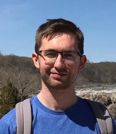 Young man with eyeglasses and blue t-shirt with hills and blue sky behind him.