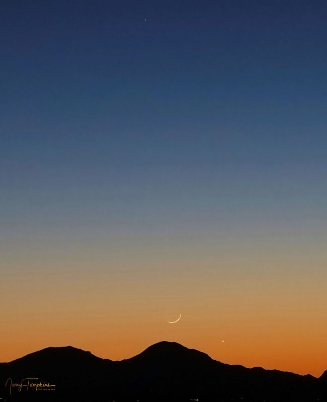 A colorful sunset with a thin crescent moon, right over a mountainous silhouette, together with two bright planets.