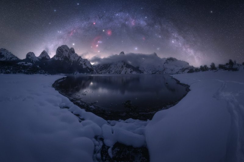 Arch of Milky Way over lake surrounded by snow; pointy mountains in distance.
