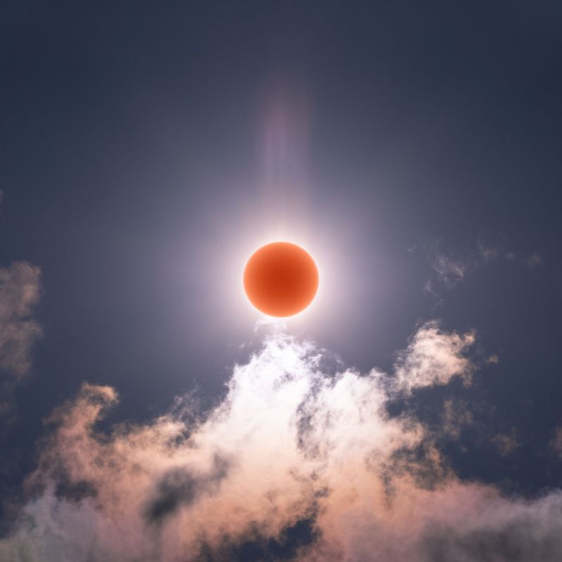 The sun imaged as a red orb in the center with rays emanating from it and lit up clouds below. 