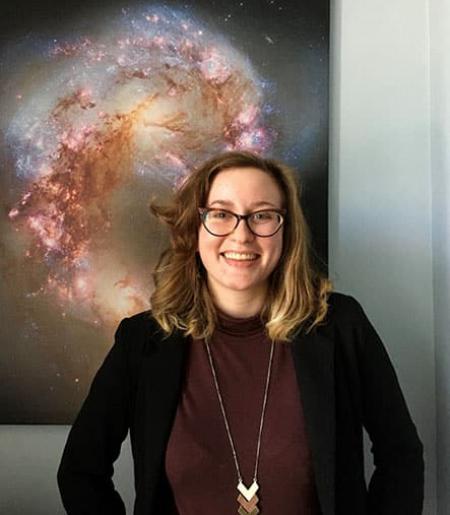 Young woman with glasses and medium-length brown hair in front of galaxy photo.