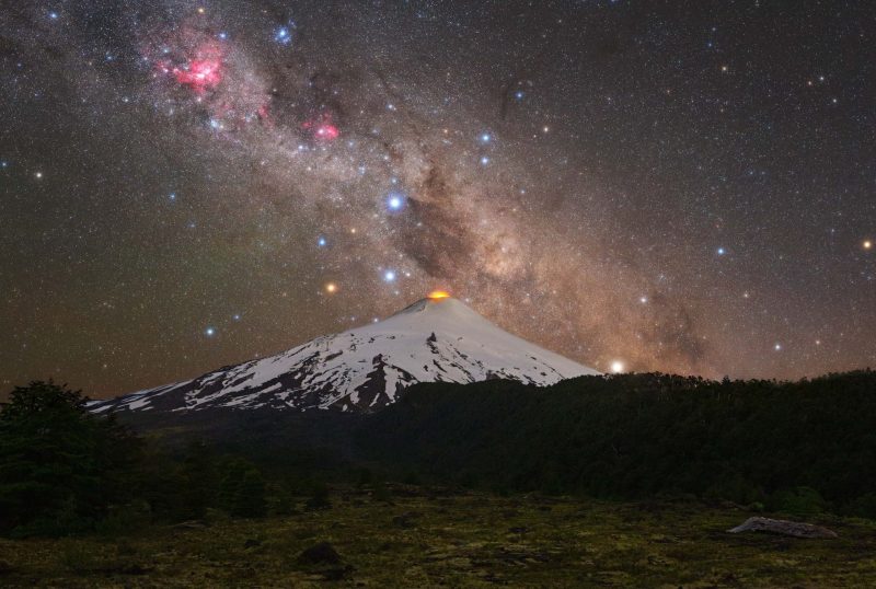 Snowcapped low conical volcano with orange lava glow and scattered stars and Milky Way above.