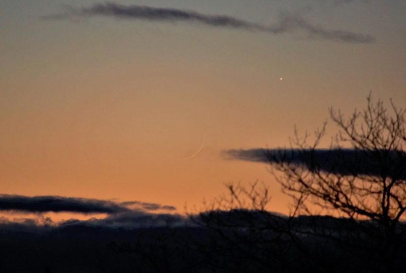 Extremely thin crescent barely visible against orange sky, with bright planet Venus.