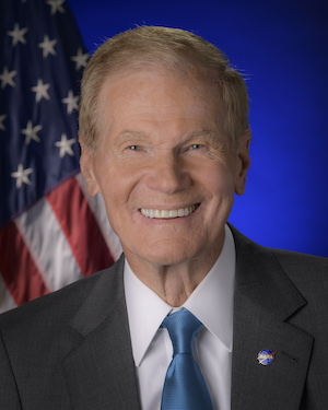 Smiling man in suit and tie with American flag behind him.