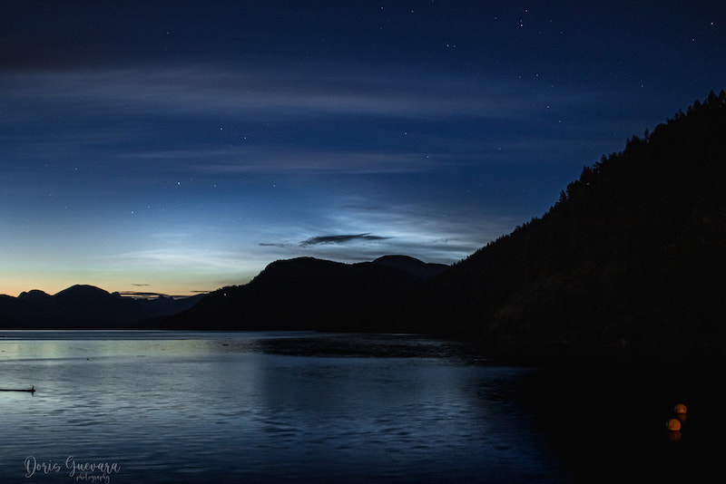 Light clouds streak across a dark blue scene with a river below and mountain silhouettes that divide the water from the sky.