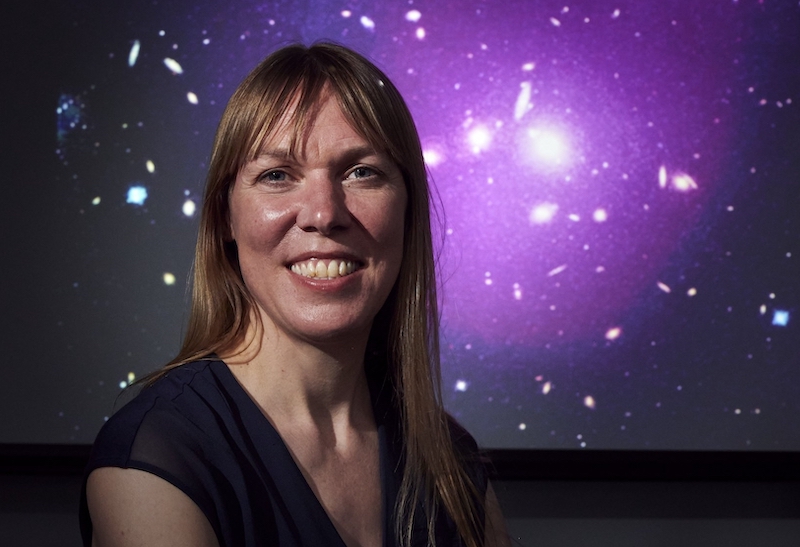 A white woman smiles in front of a purple backdrop with white dots, or stars.