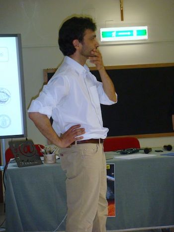 Standing man with one hand on his chin and one on his hip, with table and blackboard behind him.