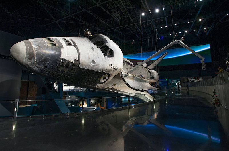 Final shuttle mission featured white space shuttle Atlantis, now on display. Image shows a black-tipped nose in a dark room with the glow of a blue Earth displayed behind it.