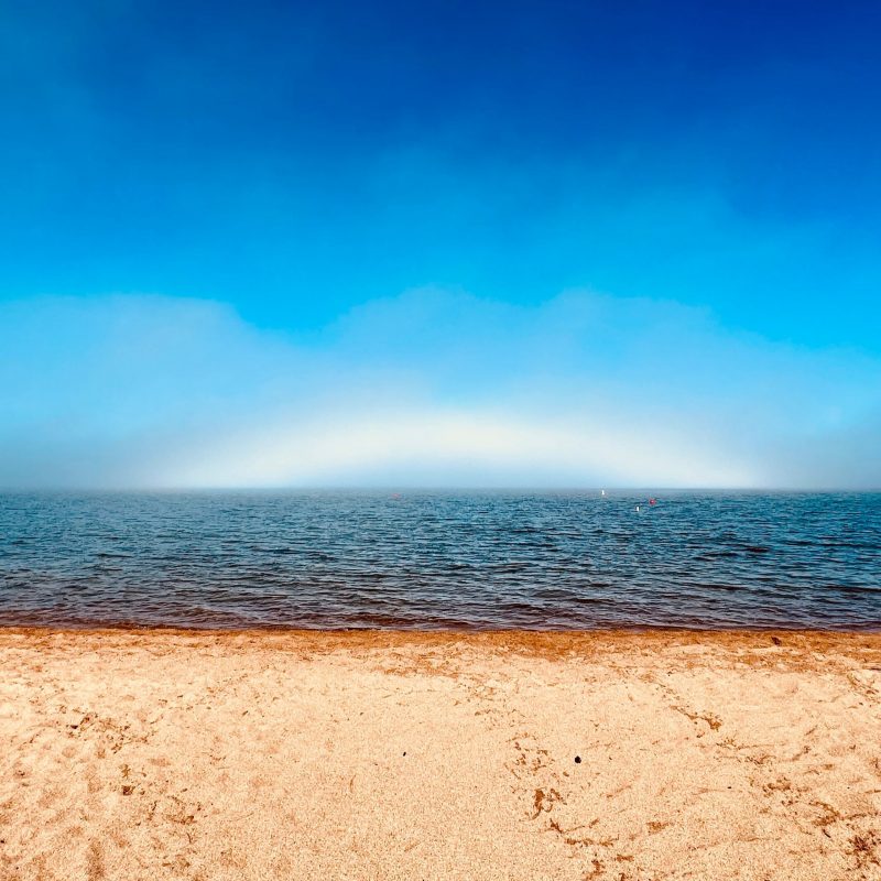 Beach and water with very low, fuzzy white cloudy arc at horizon and blue sky above.