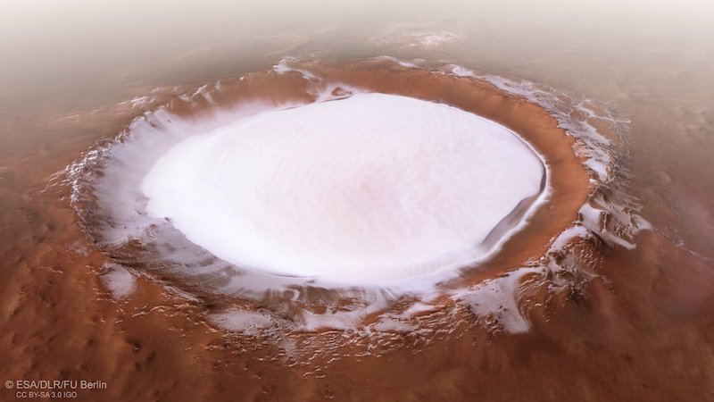 Mars ice: Oblique view of large, ice-filled crater with raised rim in landscape of red rock.