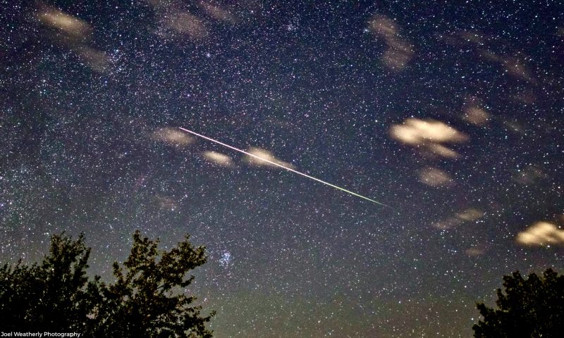 A few clouds and a thin, long, very bright meteor trail streaking across center of starry sky.