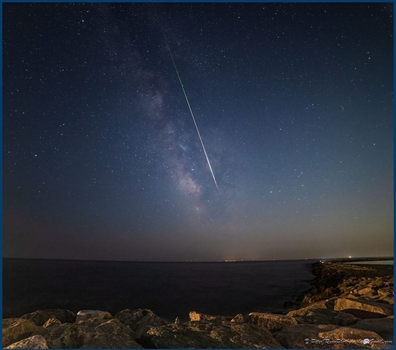 Panoramic view of the night sky with the Milky Way and a long, thin glowing white streak.