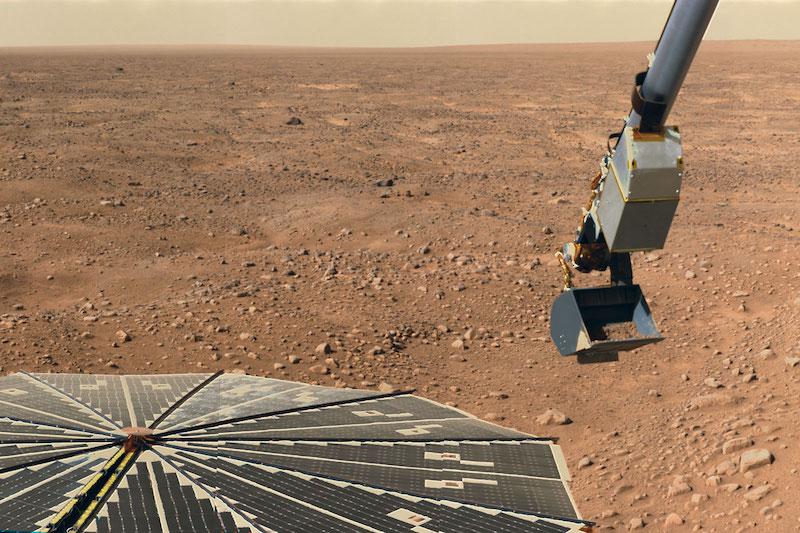 Flat panel and robotic arm with scoop and reddish rocky terrain in background.