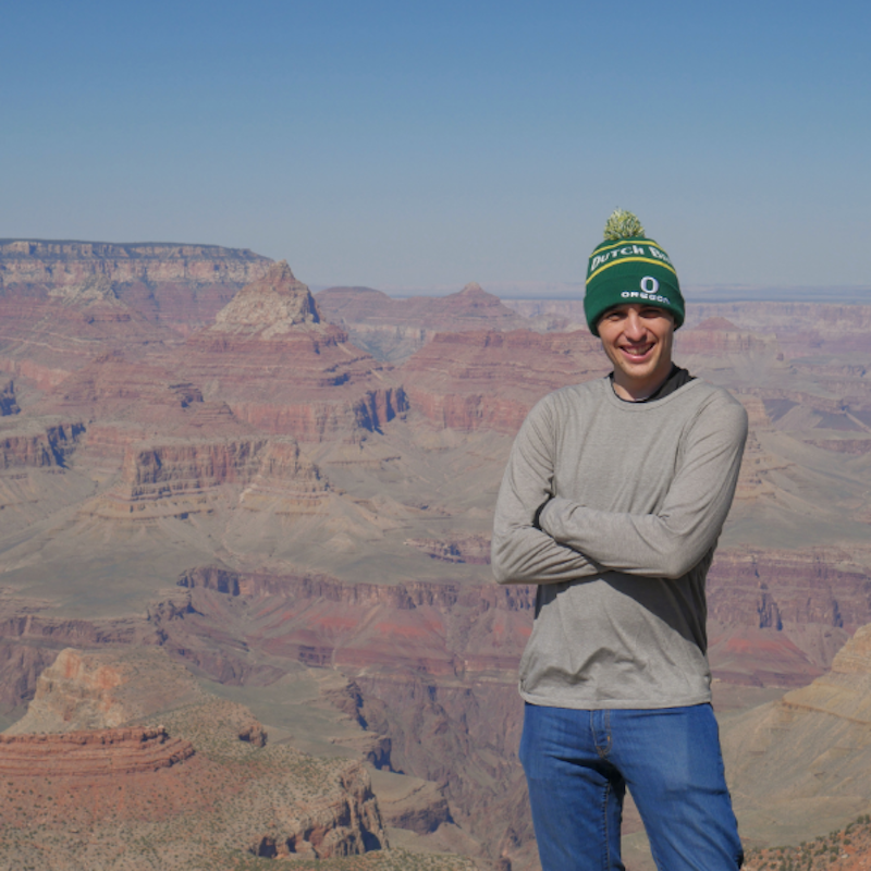 Man in knit cap, long sleeved sweater and jeans, with his arms folded, standing in front of a canyon.