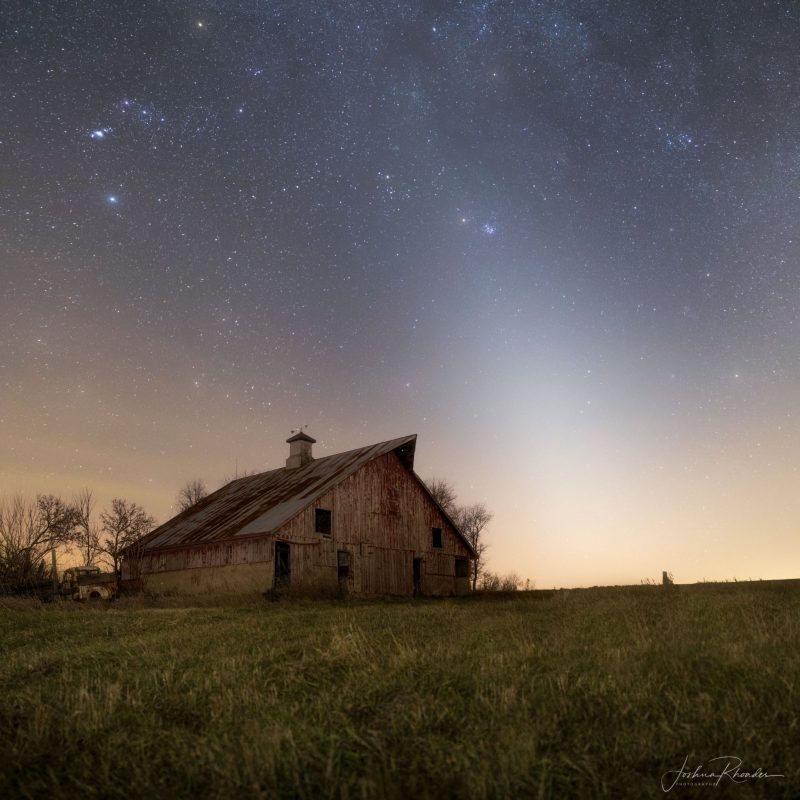 A faint triangle of light sloping toward the upper left in a starry sky over an old barn.