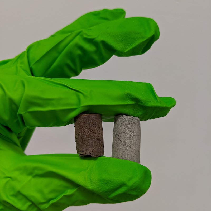 Fingers in bright green plastic glove hold small concrete cylinders, one reddish and the other gray.