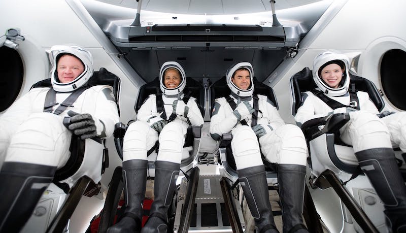 Inspiration4: 2 men and 2 women all in white spacesuits strapped into seats inside a small cabin with portholes.