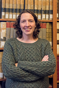 Woman in sweater standing with arms crossed. Smiling, dark curly hair to shoulders.