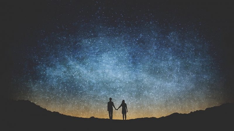 Standing couple in silhouette, holding hands under a vast starry sky.