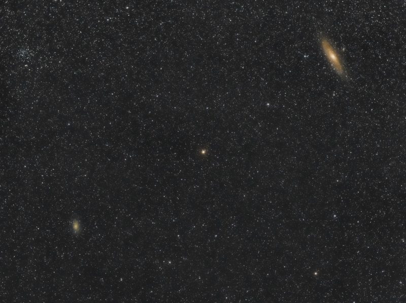 Andromeda galaxy shown as a large glowing oblong galaxy in the upper right corner, reddish star at center, hazy patch bottom left.