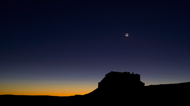 Sunset colors with crescent moon and earthshine above black silhouette of butte.