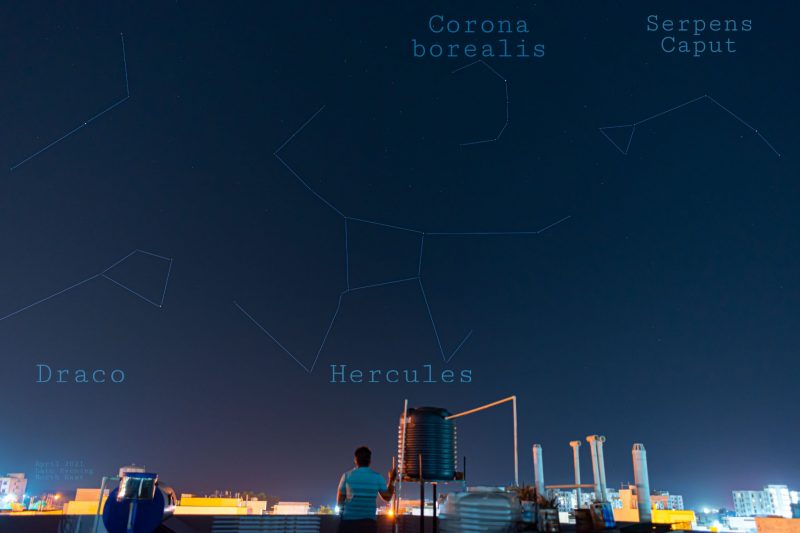 Man on rooftop of city with constellations including Hercules outlined in night sky.