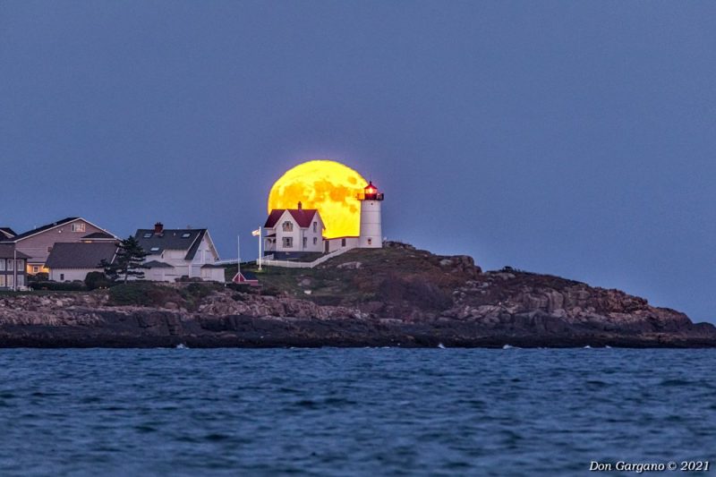 Full moon rising behind a lighthouse on a rocky coast.