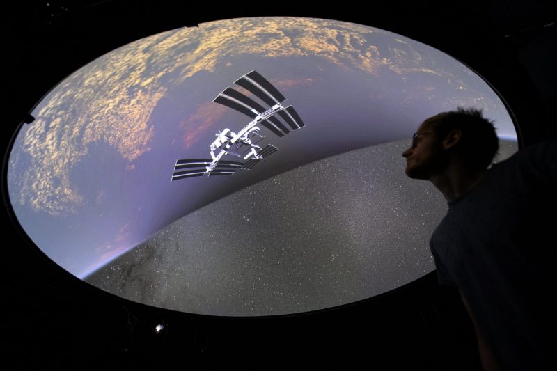 Seorang pria berdiri dalam bayang-bayang di depan gambar bulat Bumi, Stasiun Luar Angkasa Internasional, dan langit berbintang.