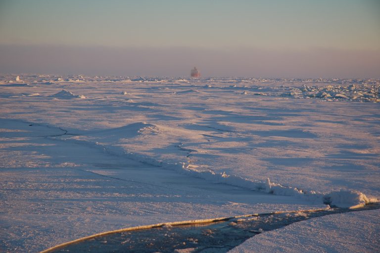 Fate of Arctic sea ice: Ice to the horizon, except a broken area showing dark water in the foreground. A distant, barely visible ship.