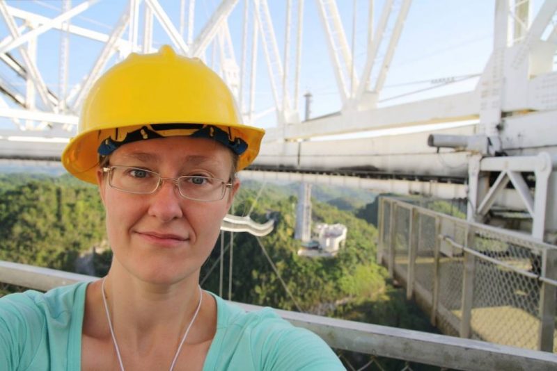 Woman with rectangular glasses in hard hat in front of large radio telescope components.