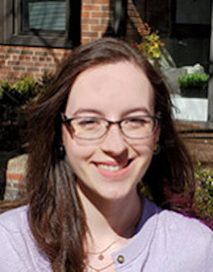 Smiling woman with eyeglasses and long dark hair in front of brick building.