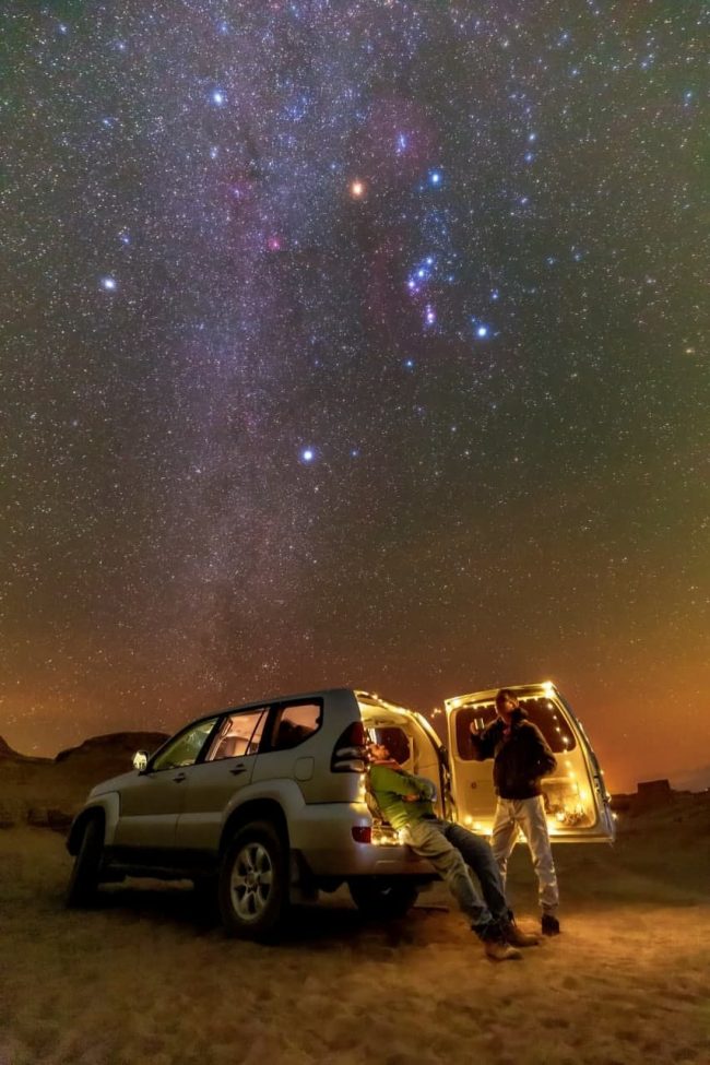 A desert landscape and SUV with people looking up at bright Orion in densely starry sky.