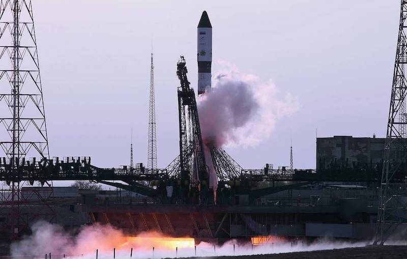 Progress 79: A white rocketship is pictured with flames and smoke beneath it on a black launch pad.