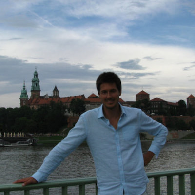 Smiling man standing on bridge by river with buildings in background.