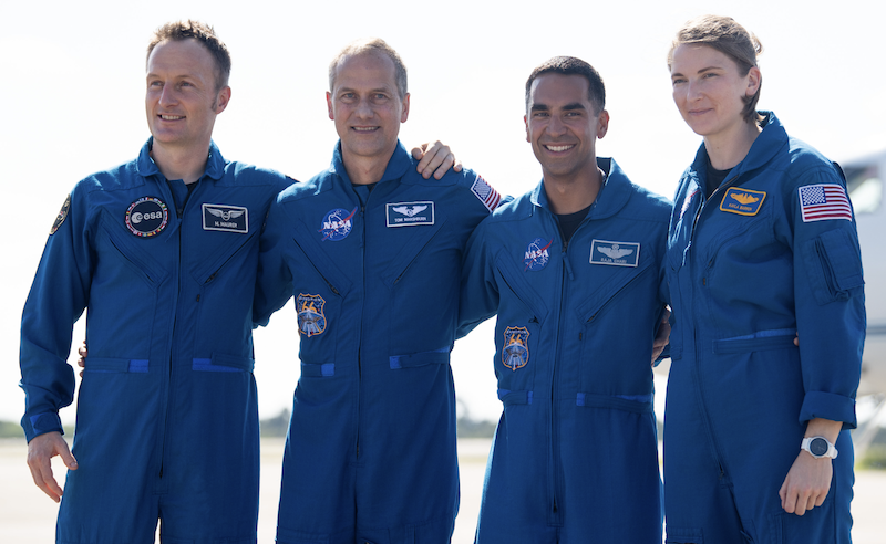 Four astronauts wearing blue jumpsuits with insignia patches stand side by side.