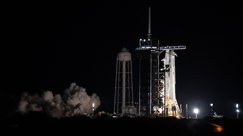 Night scene of brightly illuminated white rocket upright on a launch pad with white smoke billowing away from it.