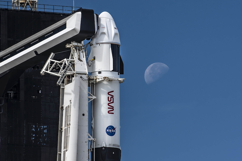 Crew-3: A white rocket is held upright by tall machinery and a faint half-lit moon shines in the blue daylight sky.