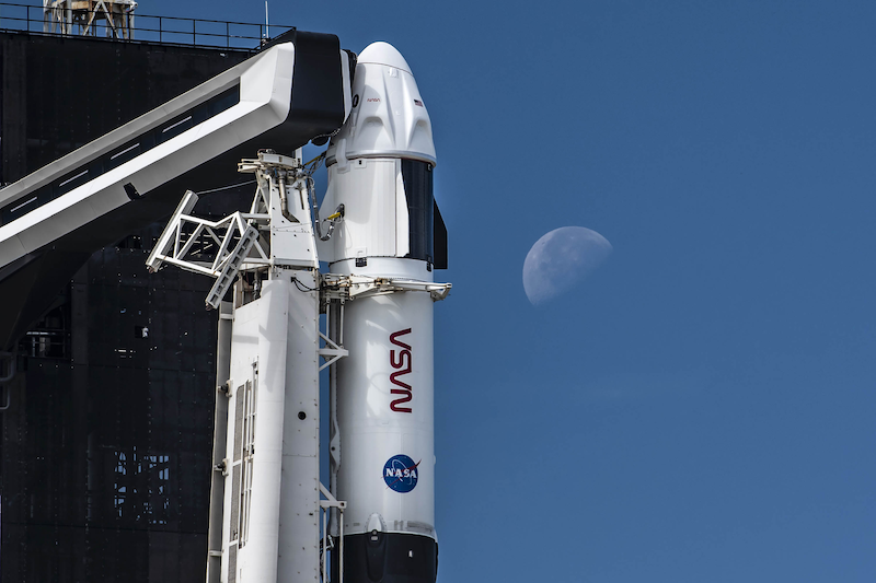 Crew-3; A white vehicle is held vertically upright with machinery while a half-lit orb is faintly seen in the background.