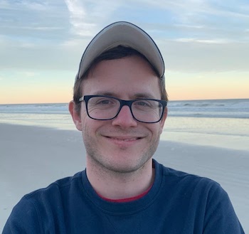 Smiling young man with glasses and ball cap and beach behind him.