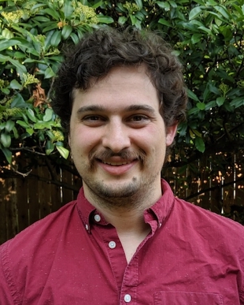 Smiling young man with goatee and beard, with trees behind him.