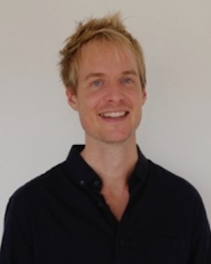 Smiling yellow-haired man with dark shirt on plain background.