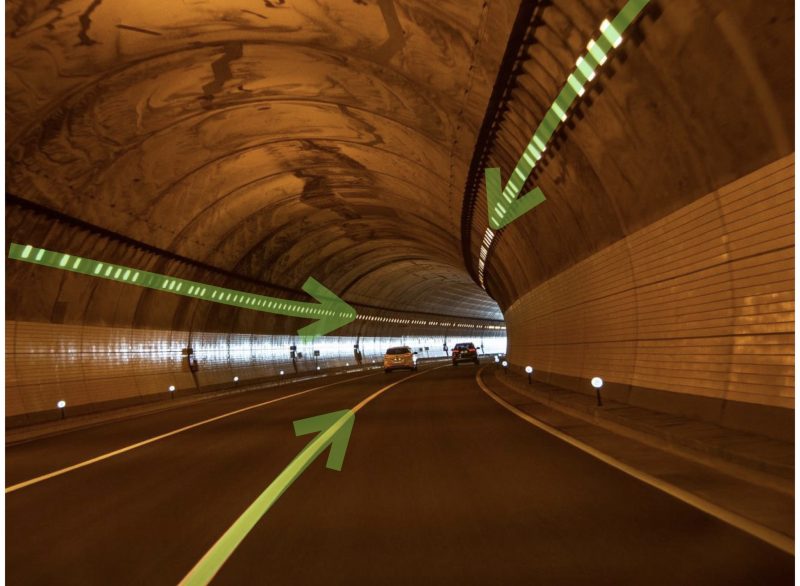 Cars in curving tunnel with green arrows pointing into the distance.
