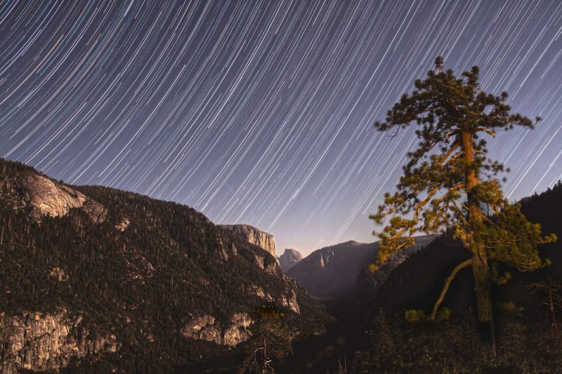Star trails above with rocky landscape below and Half Dome in the distance.
