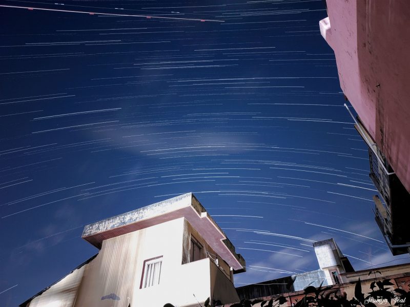 Foreground buildings with streaks of stars and one dotted plane trail in sky.