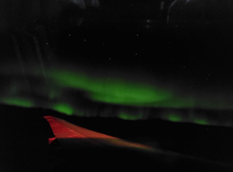 Orangish plane wing with green aurora stripe near horizon and Big Dipper behind.
