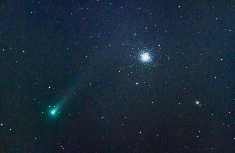 Greenish comet with tail stretching bottom left toward upper right, with cluster of lights at upper right.