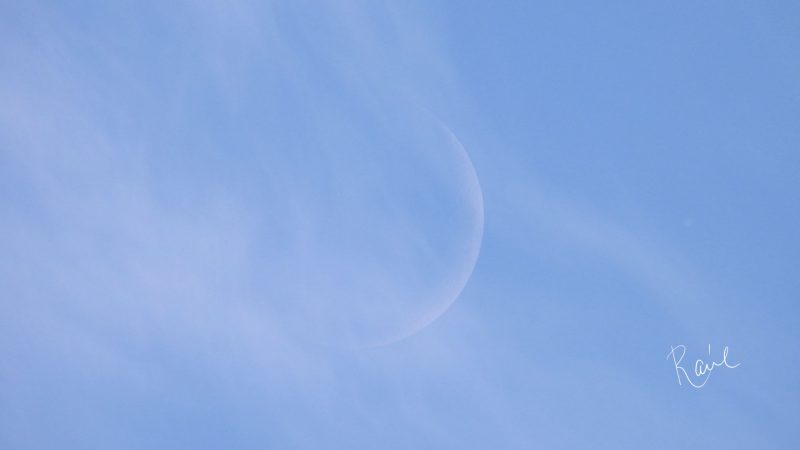 Faint but distinct crescent moon behind wispy clouds in a pale blue sky.